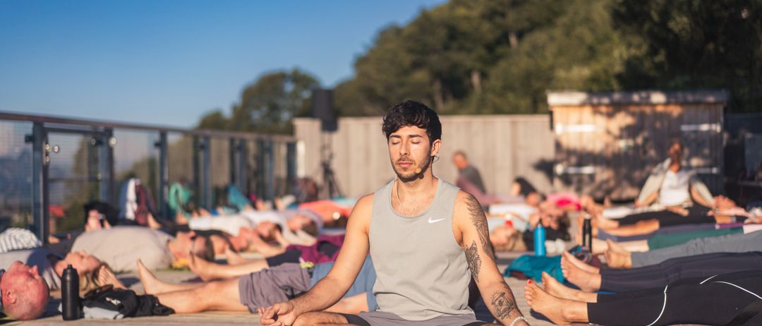 Ein Mann in einem grauen Tanktop und Shorts sitzt im Schneidersitz auf einer Yogamatte auf einer Holzterrasse, mit mehreren Personen in verschiedenen Yoga-Posen im Hintergrund.