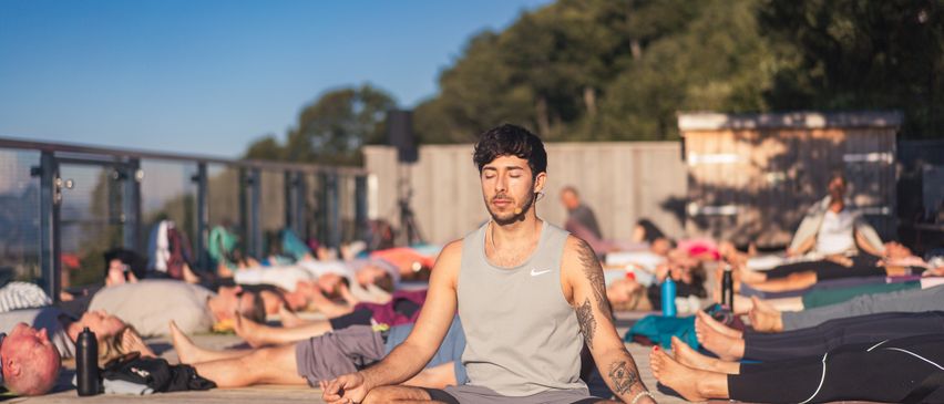 Ein Mann in einem grauen Tanktop und Shorts sitzt im Schneidersitz auf einer Yogamatte auf einer Holzterrasse, mit mehreren Personen in verschiedenen Yoga-Posen im Hintergrund.