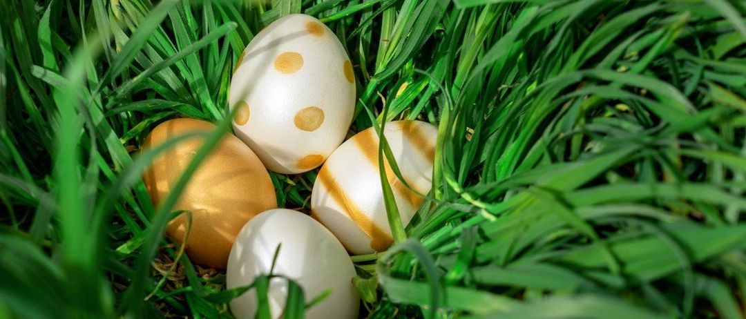 Vier dekorative Eier liegen in einem Beet aus grünem Gras. Die Eier haben verschiedene Designs, darunter goldene Punkte, goldene Streifen und schlichtes Weiß.