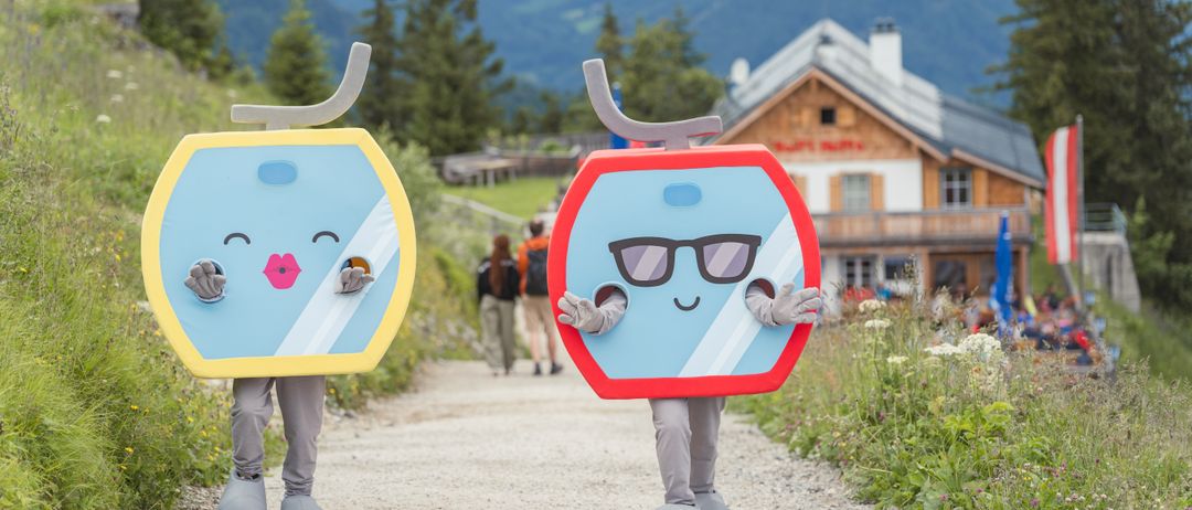 Zwei Personen in Kostümen gehen auf einem Bergweg. Eine trägt ein blaues Gondelkostüm mit Gesicht, die andere ein rotes Gondelkostüm mit Brille. Hinter ihnen gehen zwei Personen mit Rucksäcken.