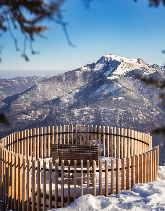 Eine Bank, umgeben von einem Holzzaun, steht auf einem verschneiten Berg mit blauem Himmel darüber.