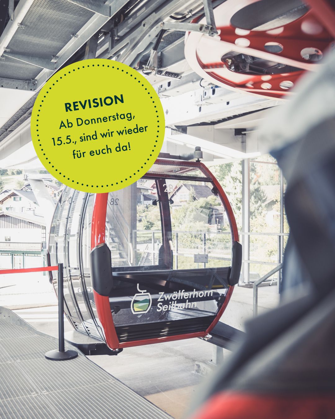 Eine Seilbahn ist geparkt mit einem gelben Schild, das 'Revision' und 'Ab Donnerstag, 15.5., sind wir wieder da!' sagt. Sie steht neben einer Metallstange mit einem Banner.