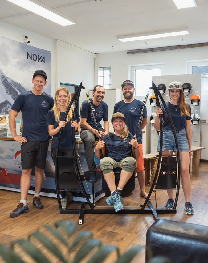 Sechs Personen stehen um eine große Trainingsmaschine herum und posieren für ein Foto. Zwei sitzen in der Maschine, während andere an deren Griffen halten. Sie tragen übereinstimmende marineblaue T-Shirts und verschiedene Outdoor-Ausrüstung.