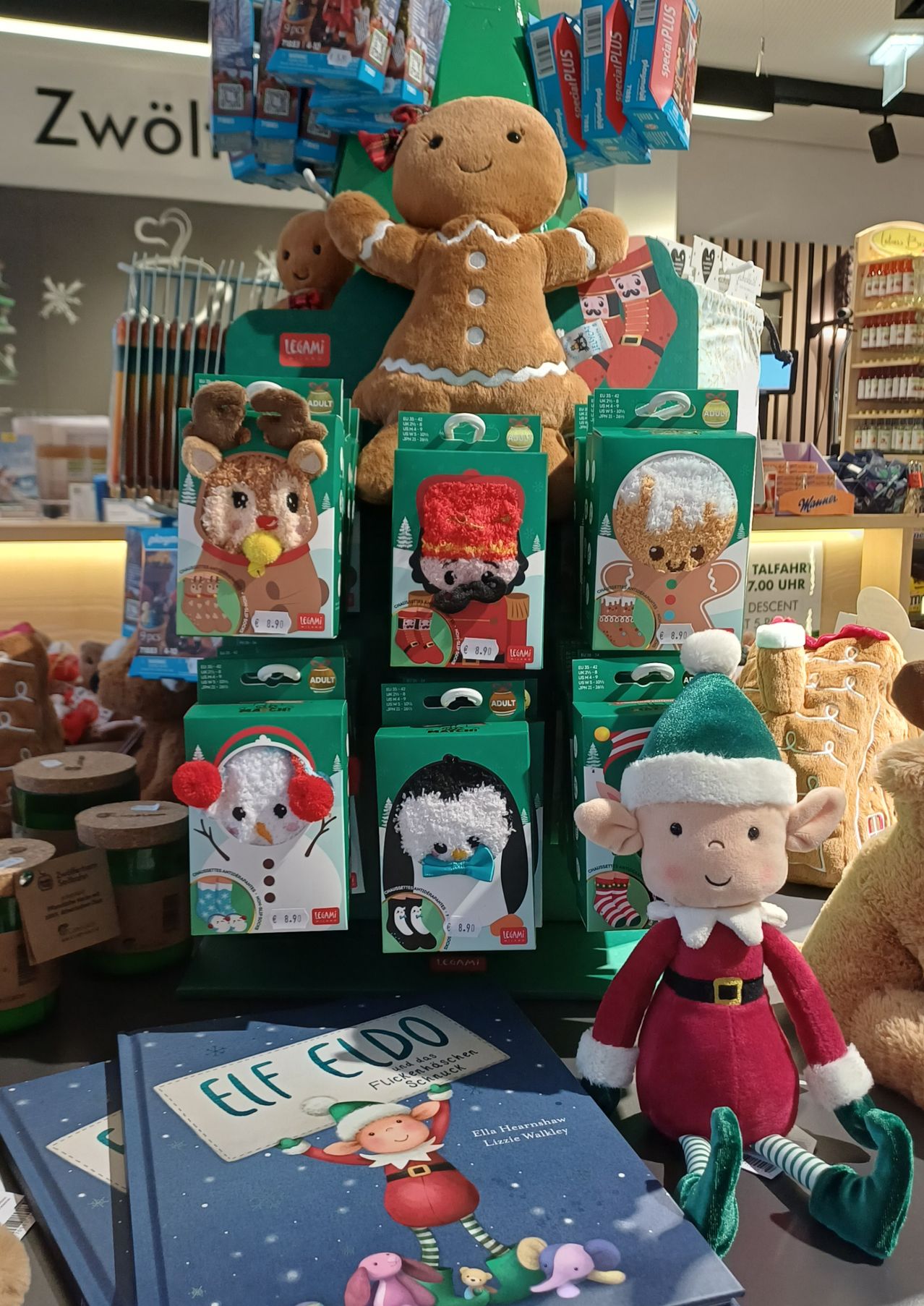 A display of Christmas stuffed toys, including gingerbread men, reindeer, and elves, arranged in green boxes, with other toys and products in the background.