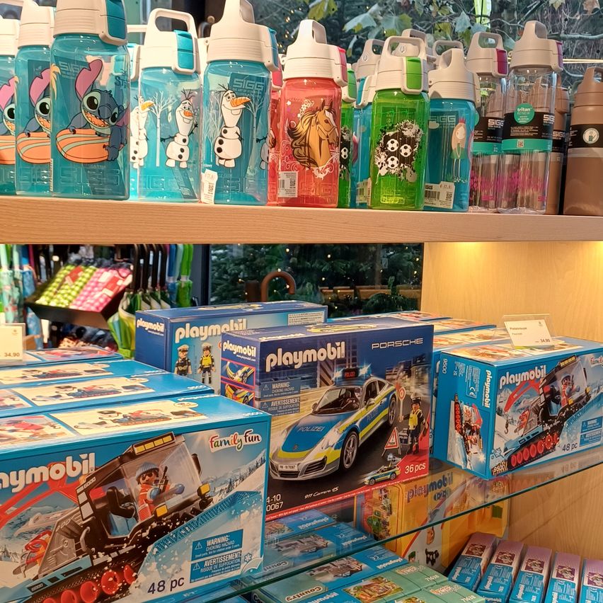 A wooden shelf with a variety of playmobil toys and water bottles. Water bottles with cartoon characters are on the top shelf. Below are boxes of playmobil toys, including a police car, a snow plow, and others.