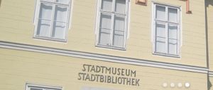 Ein Gebäude mit gelber Wand hat zwei Fenster. Unter den Fenstern befindet sich ein Schild, das Stadtmuseum Stadtbibliothek besagt.