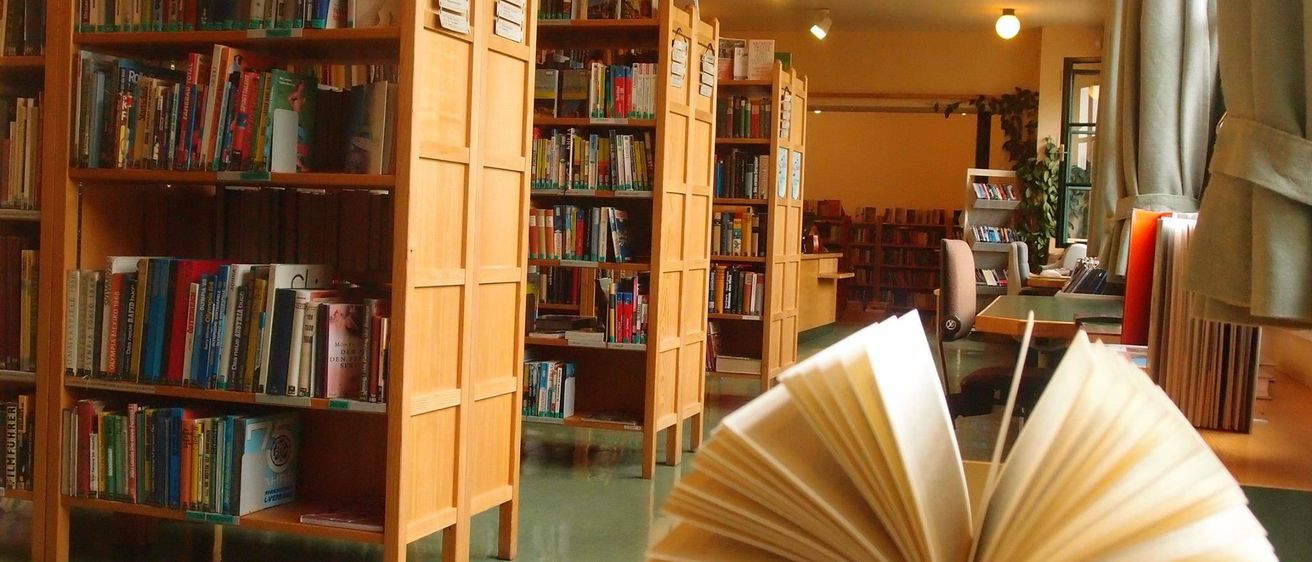 Reihen von Holzregalen sind mit Büchern gefüllt. Mehrere Lampen hängen von der Decke. Ein offenes Buch liegt auf einem Tisch.