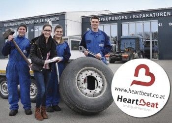 Vier Personen in blauen Overalls stehen um einen großen Reifen vor einer Garage mit Fahrzeugen im Inneren. Einer hält einen Vorschlaghammer, einer eine Schraube und einer ein Papier. Ein rundes Logo mit dem Text 'www.heartbeat.co.at' und 'Heartbeat' ist unten rechts.