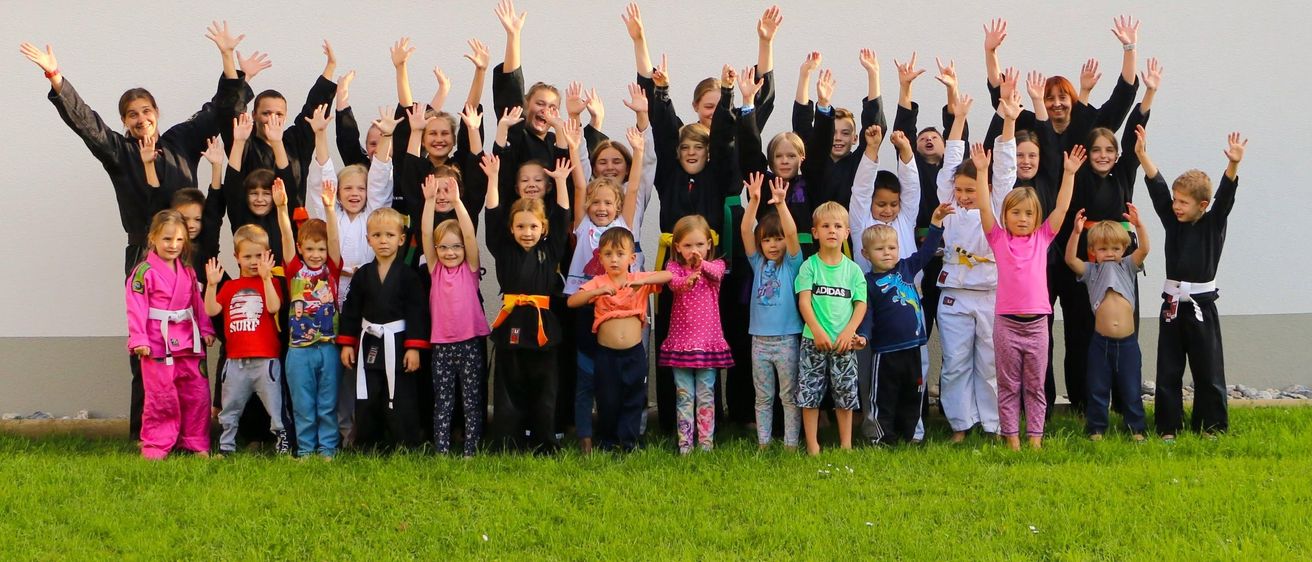 Eine Gruppe von Kindern, einige in Kampfsportuniformen, steht in einer Reihe und hebt ihre Hände, wahrscheinlich für ein Foto.