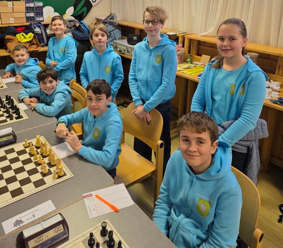 Eine Gruppe von Kindern in blauen Hoodies spielt Schach in einem Klassenzimmer mit Holzstühlen und einer Uhr auf dem Tisch.