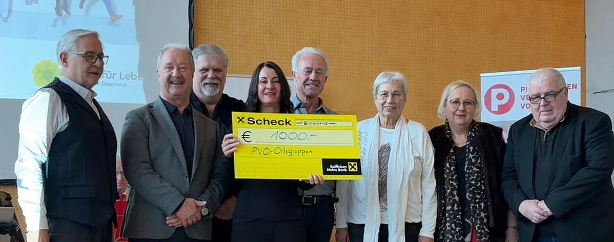 Eine Gruppe von Menschen steht eng beieinander und lächelt. Eine Frau in der Mitte hält einen gelben Scheck über 1000 Euro. Hinter ihnen zeigt eine Präsentationsfolie das Wort 'Leben'.