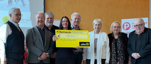 Eine Gruppe von Menschen steht eng beieinander und lächelt. Eine Frau in der Mitte hält einen gelben Scheck über 1000 Euro. Hinter ihnen zeigt eine Präsentationsfolie das Wort 'Leben'.