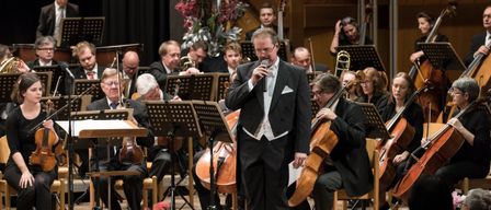 Ein Mann singt vor einem Orchester, mit Musikern, die Violinen, Celli und Trompeten spielen. Das Orchester sitzt auf Stühlen, mit Notenständern davor. Das Publikum sitzt vor der Bühne und verfolgt die Aufführung.