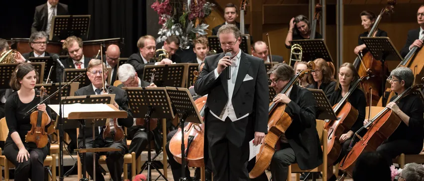 A man is singing in front of an orchestra, with musicians playing violins, cellos, and trumpets. The orchestra is seated on chairs, with music stands in front. The audience is seated in front of the stage, watching the performance.