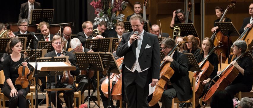 Ein Mann singt vor einem Orchester, mit Musikern, die Violinen, Celli und Trompeten spielen. Das Orchester sitzt auf Stühlen, mit Notenständern davor. Das Publikum sitzt vor der Bühne und verfolgt die Aufführung.