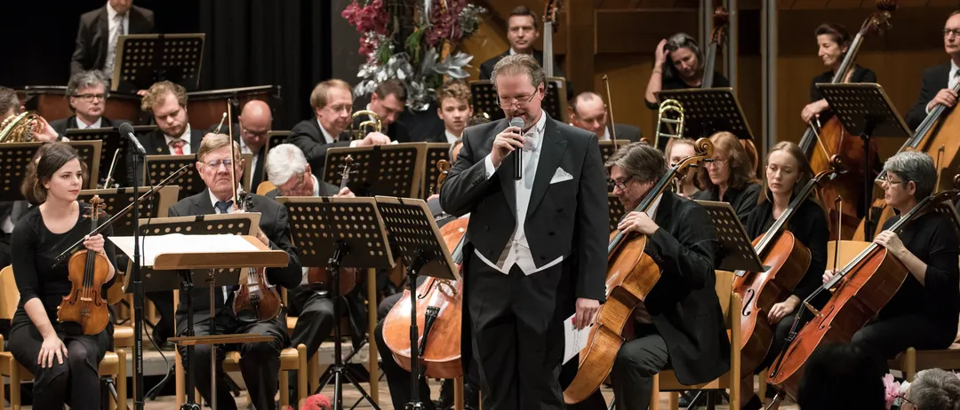 A man is singing in front of an orchestra, with musicians playing violins, cellos, and trumpets. The orchestra is seated on chairs, with music stands in front. The audience is seated in front of the stage, watching the performance.