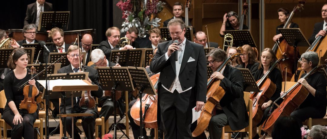 Ein Mann singt vor einem Orchester, mit Musikern, die Violinen, Celli und Trompeten spielen. Das Orchester sitzt auf Stühlen, mit Notenständern davor. Das Publikum sitzt vor der Bühne und verfolgt die Aufführung.