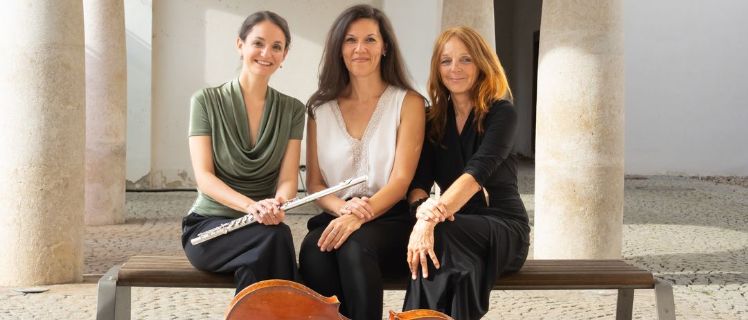 Drei Frauen sitzen auf einer Bank, eine hält eine Flöte, eine spielt Cello und eine andere trägt ein schwarzes Shirt, lächelt und posiert für ein Foto.