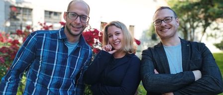 Drei lächelnde Personen stehen zusammen vor einem Gebäude mit weißer Wand, umgeben von roten Blumen und Bäumen. Ein Mann links trägt ein blaues kariertes Hemd, ein anderer Mann rechts trägt einen schwarzen Mantel, und eine Frau in der Mitte trägt einen schwarzen Blazer.