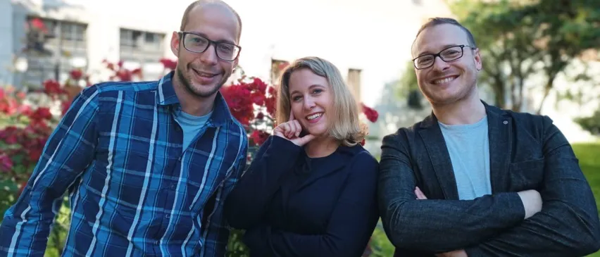 Three smiling individuals stand together in front of a building with a white wall, surrounded by red flowers and trees. A man on the left wears a blue plaid shirt, another man on the right wears a black jacket, and a woman in the middle wears a black blazer.