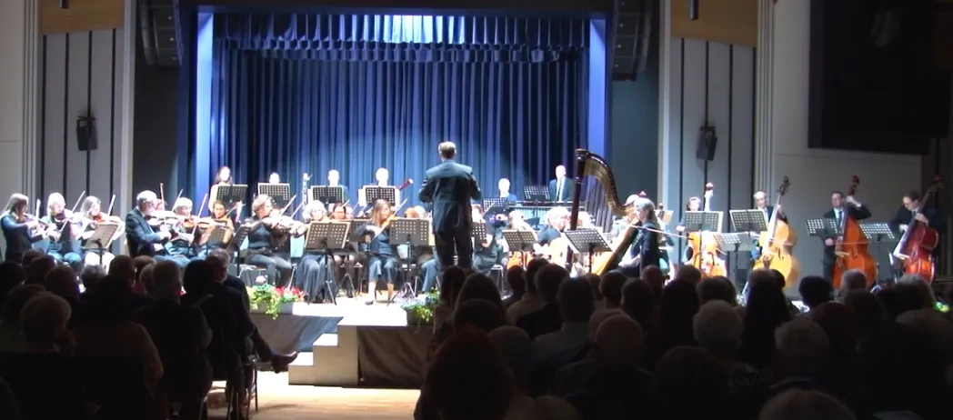 An orchestra is performing on a stage with blue curtains. The audience is seated in front, watching. The orchestra members are playing various instruments.