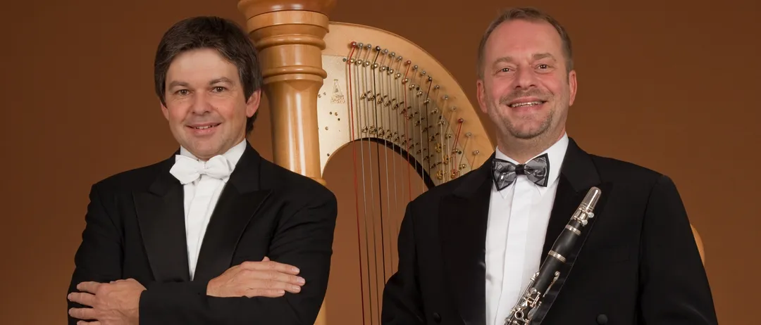 Two men in formal attire stand next to each other. One holds a clarinet, and the other stands next to a harp. They both smile and appear to be posing for a photo.
