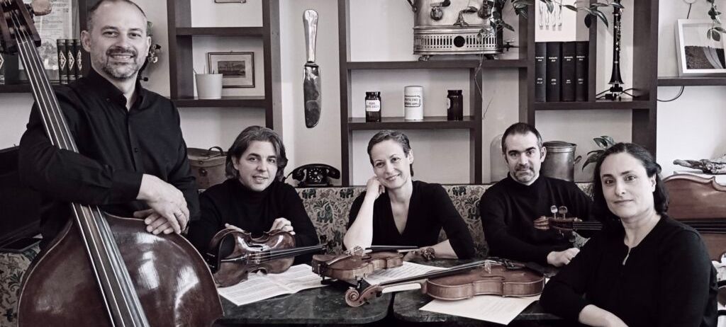 Fünf Musiker, zwei Männer und drei Frauen, posieren für ein Foto. Violinen, Cello und Notenblätter liegen auf dem Tisch vor ihnen. Regale mit verschiedenen Gegenständen stehen hinter ihnen.