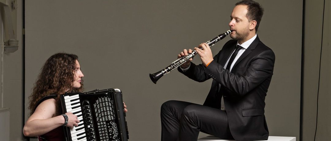 Ein Mann und eine Frau sitzen auf weißen Blöcken, formal gekleidet. Der Mann spielt Klarinette, während die Frau Akkordeon spielt. Sie befinden sich in einem Raum mit einer weißen Wand.