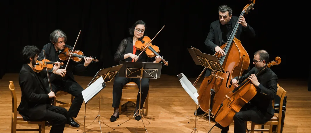 Ein Streichquartett tritt auf der Bühne auf, mit Violinen und Celli, Musiker in formeller Kleidung, sitzend auf Stühlen, beleuchtet von Bühnenlichtern, vor einem dunklen Hintergrund.