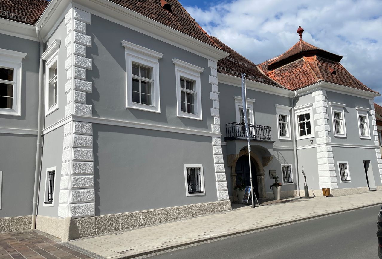 Ein graues Gebäude mit einem braunen Dach steht an der Straße. Es gibt mehrere Fenster mit weißen Rahmen. Der Eingang ist eine schwarze Tür mit einem kleinen Balkon.