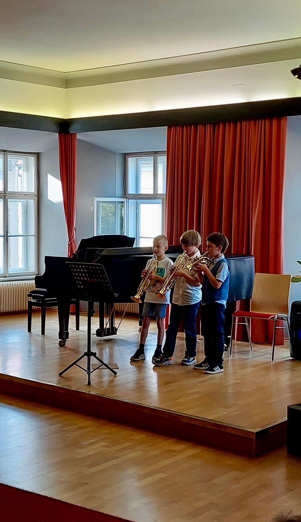 Drei Jungen spielen Trompeten auf einer Bühne in einem Musikzimmer mit Klavier und roten Vorhängen.