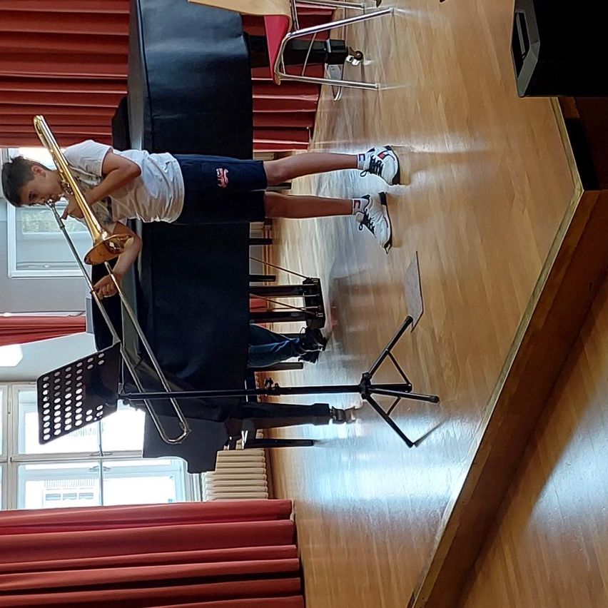 Ein junger Junge spielt Posaune auf einer Bühne mit einem Klavier im Hintergrund. Die Bühne hat einen Holzboden und rote Vorhänge.