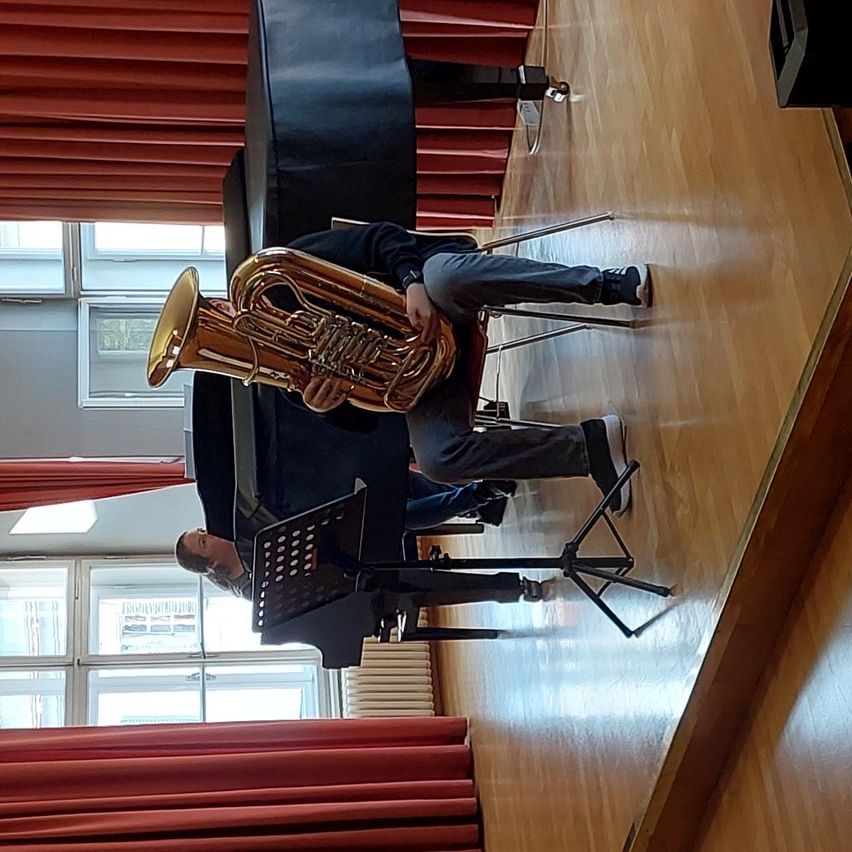 Ein Musiker spielt eine Tuba, während er auf einem Stuhl auf der Bühne sitzt. Eine weitere Person sitzt hinter ihm an einem Klavier. Die Bühne ist mit roten Vorhängen und einem Holzboden geschmückt.