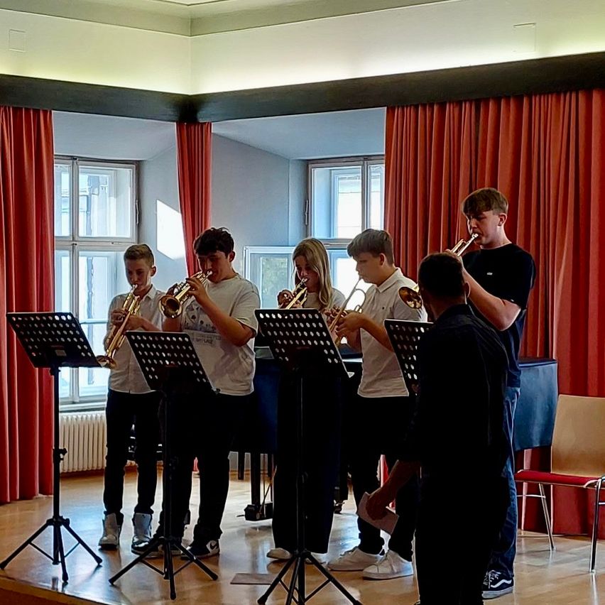 Eine Gruppe junger Musiker spielt Trompeten auf einer Bühne mit Notenständern, einem Klavier und einem roten Vorhang.