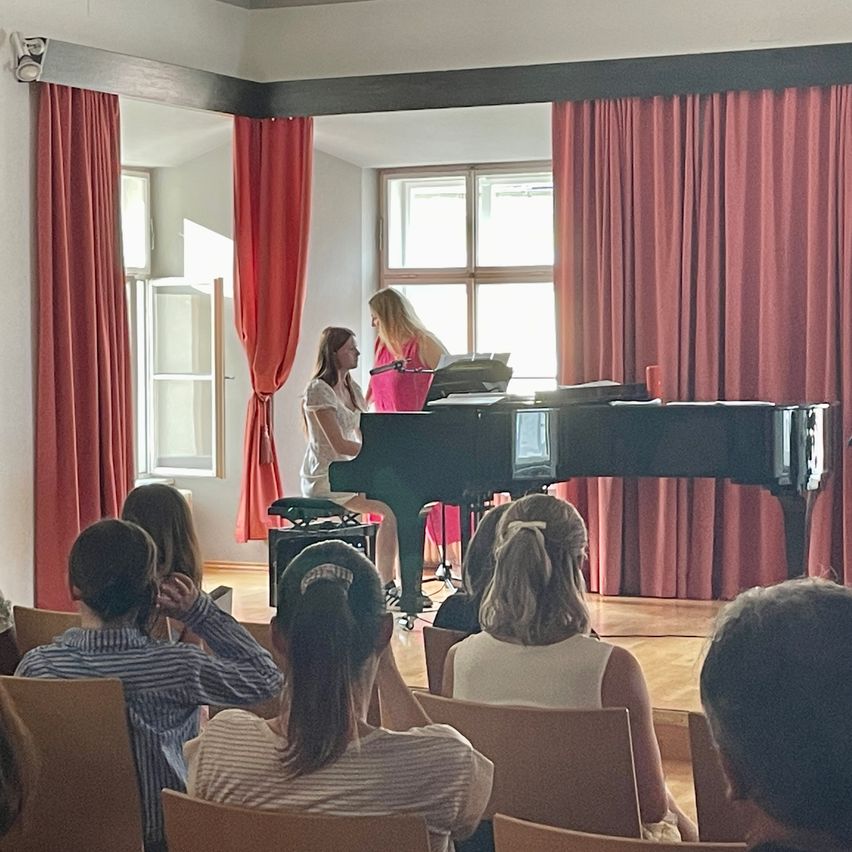 Zwei Frauen spielen ein Klavier mit einem Mikrofon vor einem roten Vorhang, den eine kleine Menge beobachtet.