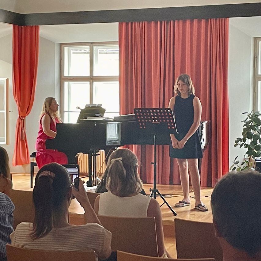 Eine Frau spielt Klavier, während eine andere Frau vor einem Notenständer steht. Eine Frau hält den Moment mit ihrem Handy fest. Sie befinden sich auf einer Bühne mit einem roten Vorhang im Hintergrund. Menschen sitzen in Stühlen und beobachten die Aufführung.