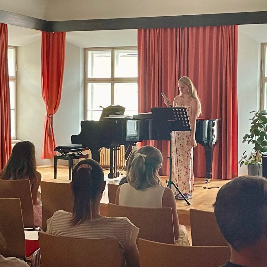 Eine junge Frau singt und hält ein Mikrofon vor einem Klavier. Mehrere Personen sitzen vor ihr auf Stühlen. Rote Vorhänge hängen im Hintergrund.