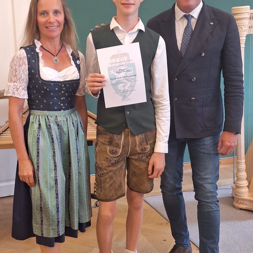Eine Frau und ein Mann in traditioneller bayerischer Kleidung stehen neben einem jungen Jungen, der ein Zertifikat in der Hand hält, alle posieren für ein Foto im Innenbereich.