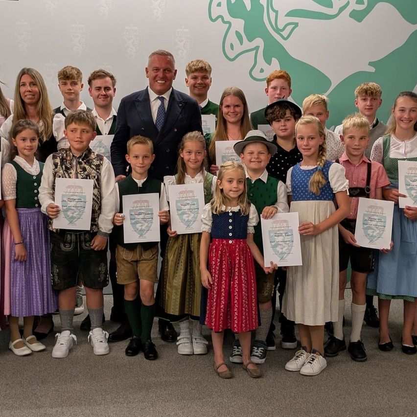 Eine Gruppe von Kindern und Erwachsenen, gekleidet in traditioneller Kleidung, posiert für ein Foto mit einem lächelnden Mann in Anzug und Krawatte. Sie halten Urkunden in den Händen.