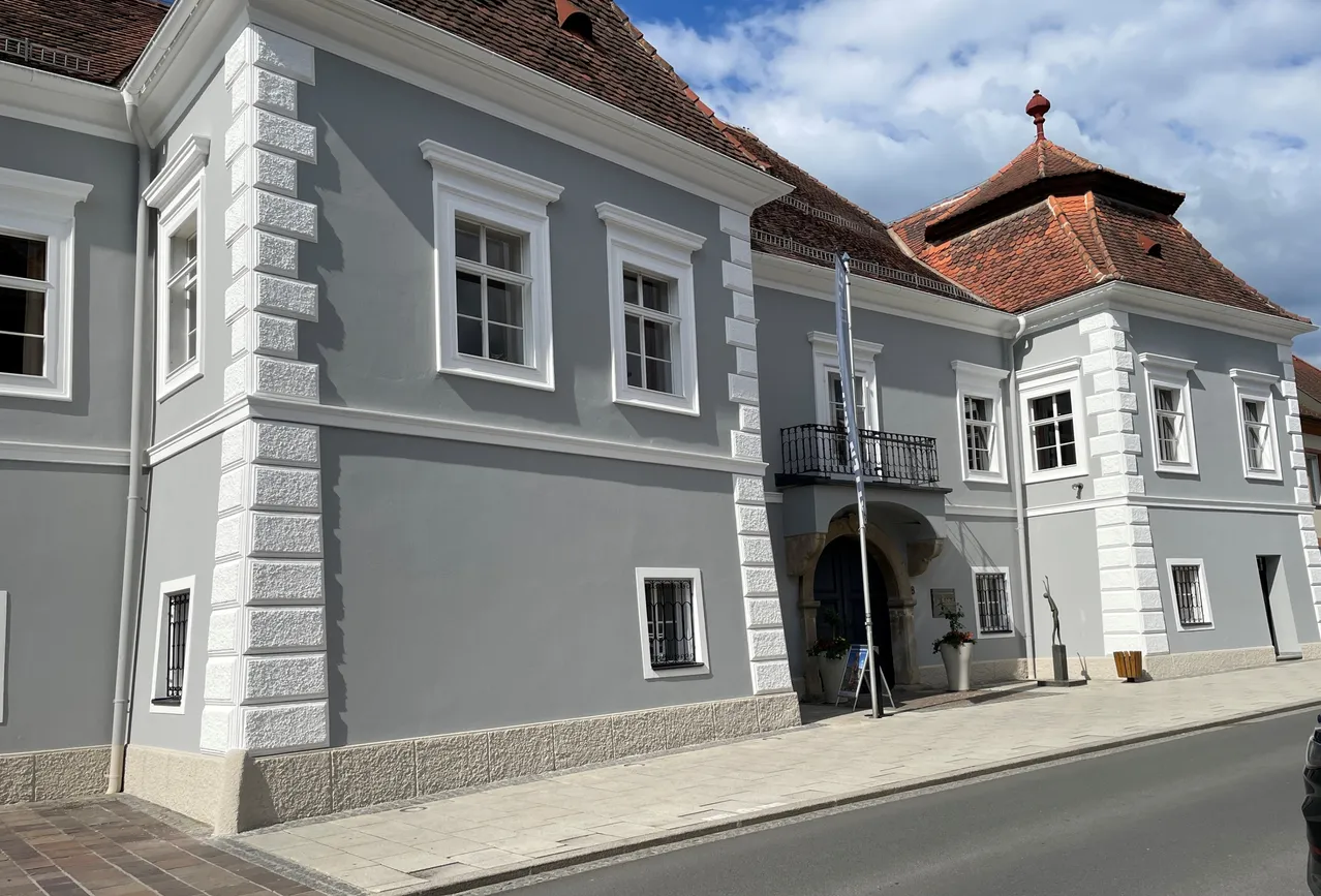 Ein graues Gebäude mit weißen Fenstern und Verkleidungen, einem braunen Dach und einem Balkon mit einem Fahnenmast davor.