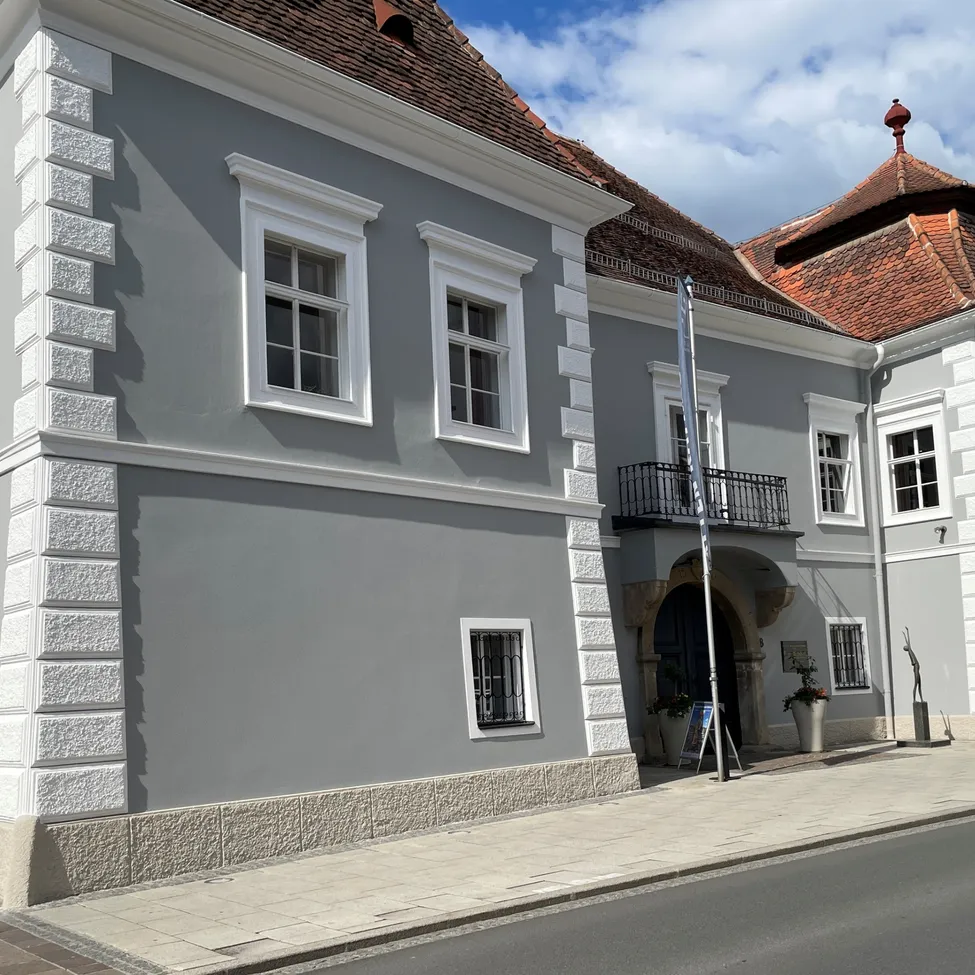 Ein graues Gebäude mit weißen Fenstern und Verkleidungen, einem braunen Dach und einem Balkon mit einem Fahnenmast davor.
