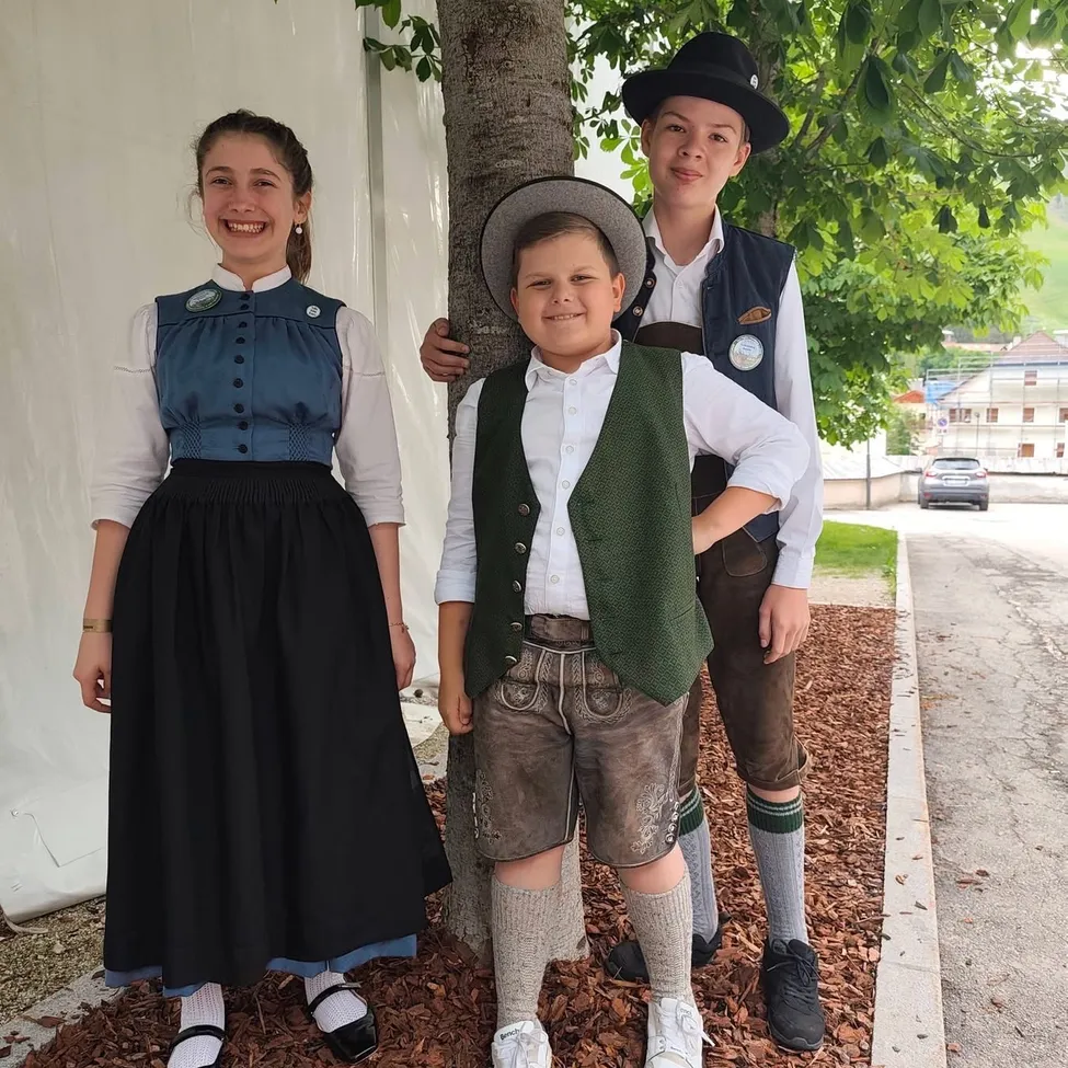 Drei Kinder in traditioneller bayerischer Kleidung stehen neben einem Baum und lächeln für die Kamera.