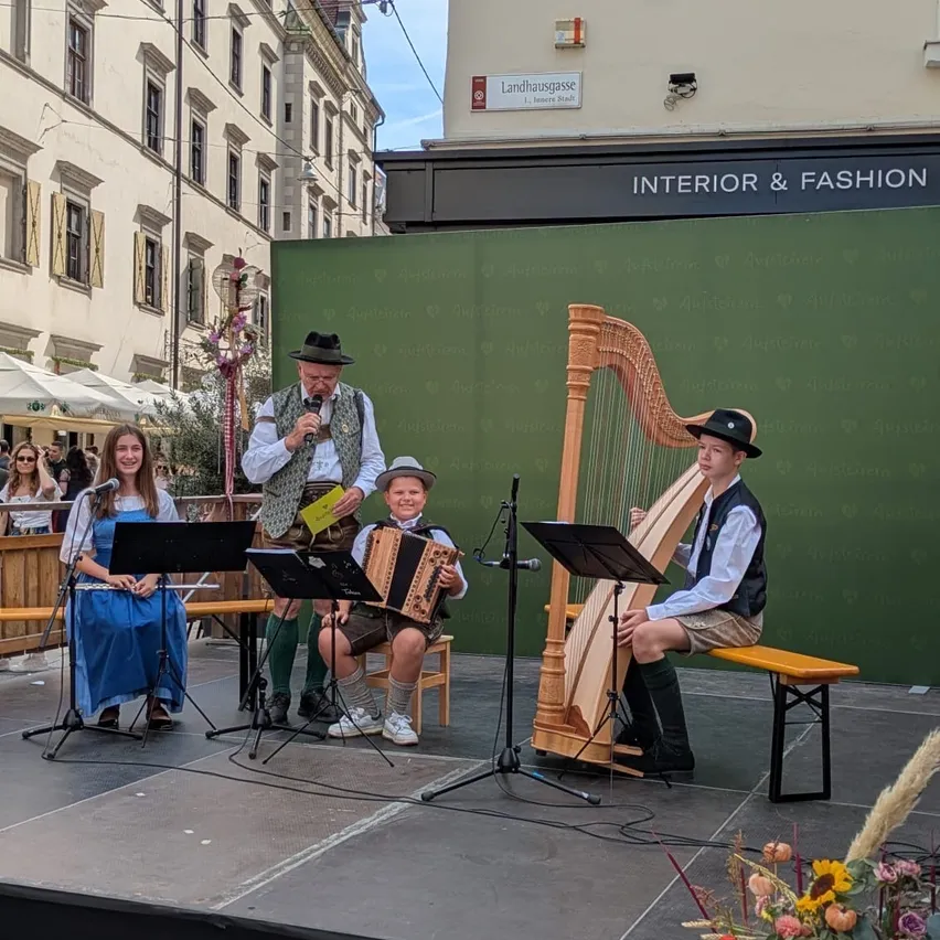 Eine Gruppe von Musikern tritt auf einer Bühne mit einem Hintergrund aus einer grünen Wand und einem Schild für Interior & Fashion auf. Die Musiker umfassen einen Mann, der Harfe spielt, einen anderen, der Akkordeon spielt, und eine Frau, die Flöte spielt.