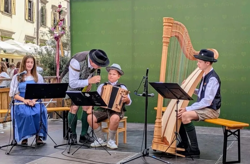 Drei Musiker spielen auf einer Bühne. Ein Mann spielt Akkordeon, ein Junge spielt Harfe und ein anderer Junge singt. Sie stehen vor einem grünen Hintergrund.