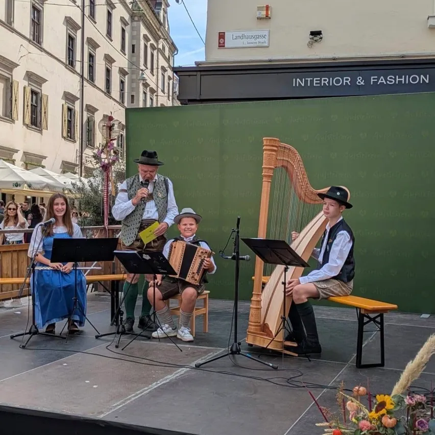 Eine Gruppe von Musikern tritt auf einer Bühne mit einem Hintergrund aus einem grünen Banner auf. Ein Mann spielt die Harfe, ein anderer spielt Akkordeon und eine Frau spielt Flöte.