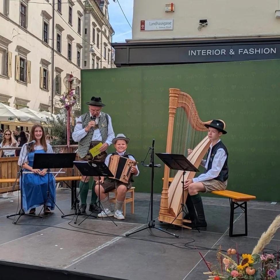 Eine Gruppe von Musikern tritt auf einer Bühne mit einem Hintergrund aus einem grünen Banner auf. Ein Mann spielt die Harfe, ein anderer spielt Akkordeon und eine Frau spielt Flöte.