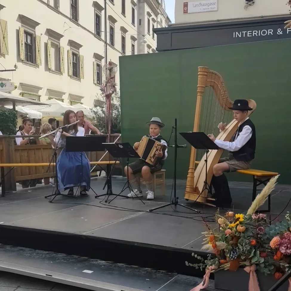 Drei Kinder spielen auf einer Bühne mit Musikinstrumenten. Ein Mädchen spielt Flöte, ein Junge Akkordeon und ein anderer Junge Harfe. Dahinter steht ein Gebäude mit grünen Wänden und einem Schild mit der Aufschrift 'Interior & F'.