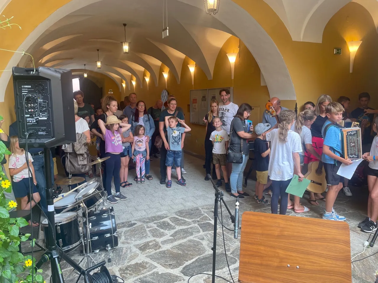 Eine Gruppe von Menschen in einem gewölbten Raum mit Instrumenten, einschließlich einer Gitarre, Trommeln und einem Lautsprecher. Einige Leute halten Papiere und schauen einen Menschen vorne an.