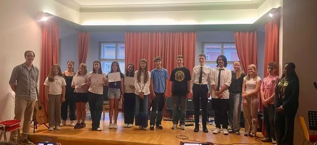 Eine Gruppe junger Menschen steht mit Zertifikaten auf der Bühne, in einem Raum mit Holzböden und roten Vorhängen. Ein Mann in einem hellblauen Shirt steht vor einer Gitarre.
