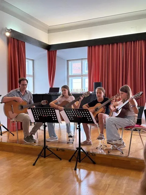 Eine Gruppe von Menschen spielt Gitarren auf einer Bühne mit Notenständern und einem Klavier im Hintergrund.